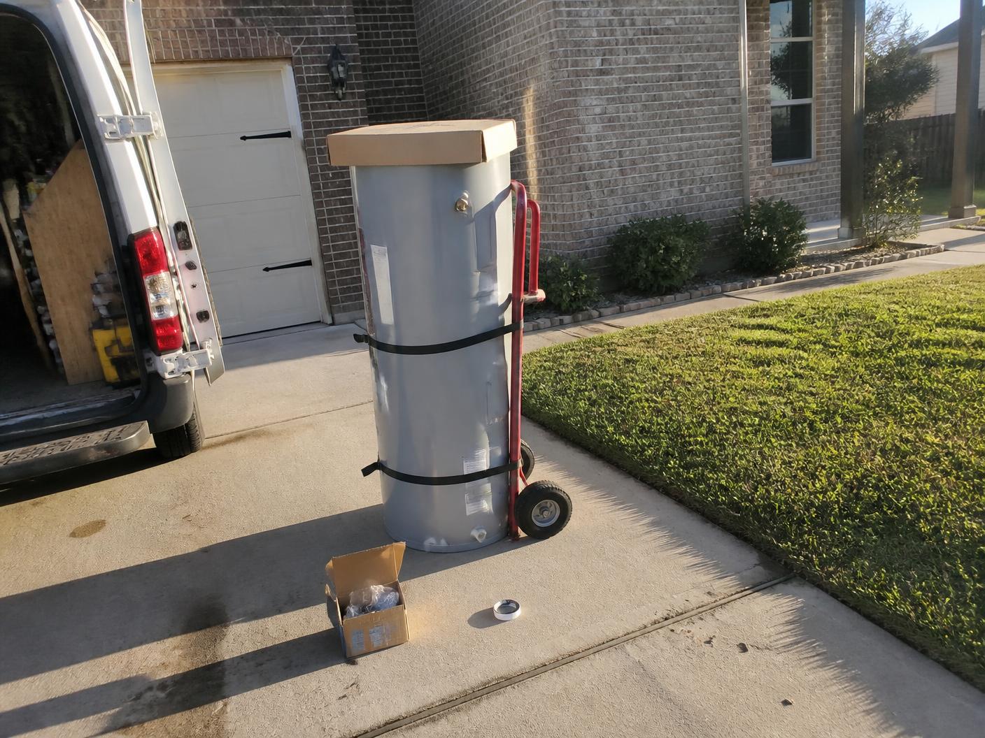 New residential water heater strapped to an appliance dolly in a suburban driveway beside an open work van
