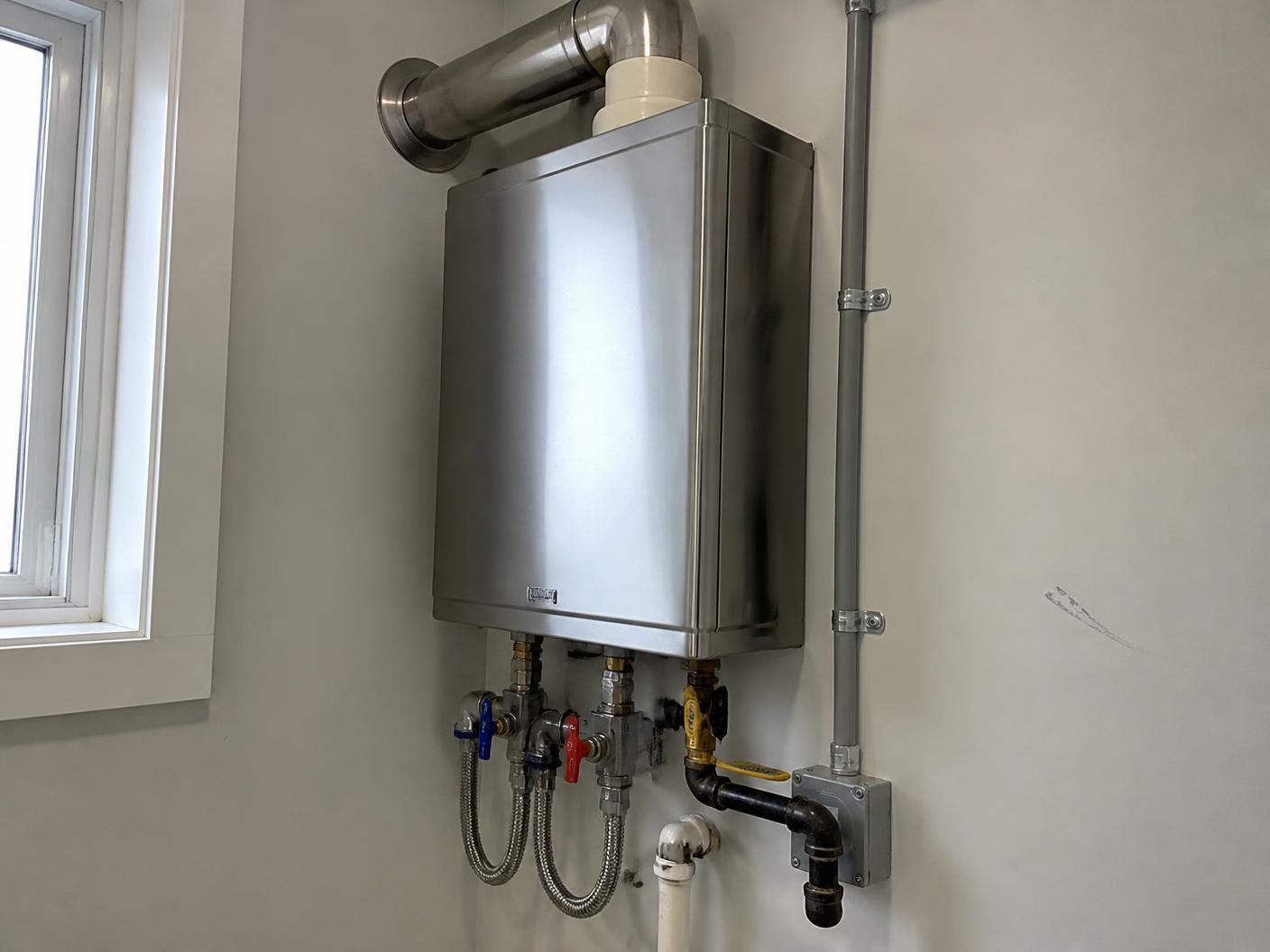 Stainless tankless water heater mounted on a utility room wall with isolation valves and vent piping
